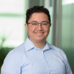 Headshot of Jake Campbell: Korean/American man with grey glasses frames, short dark hair, and a blue shirt