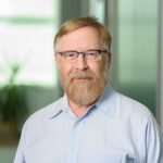 Headshot of Simon Bates: White man with short blond hair and beard, dark glasses, and blue shirt