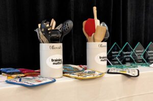 Image of a white table on a stage with various kitchen tools on top, with two containers with the words "Training matters" on the labels. 