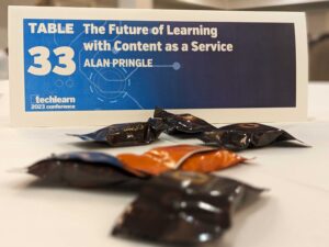 Close-up of a white table with brown and orange chocolates scattered in front of a small blue table sign that says "Table 33: The Future of Learning Content with Content as a Service, Alan Pringle."