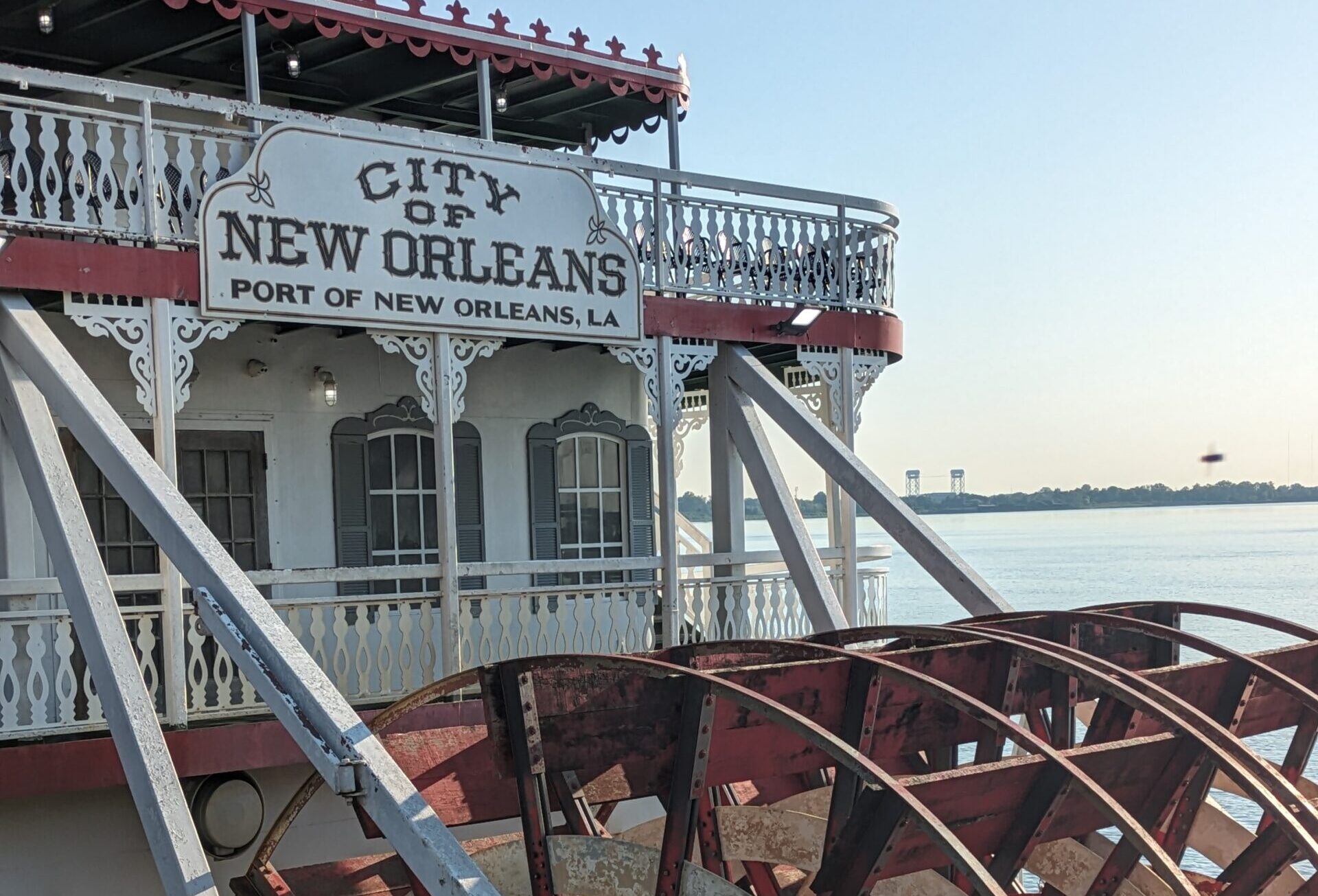 Image of a red and white boat with four large red circular paddles and a sign saying, “City of New Orleans” with water in the background.