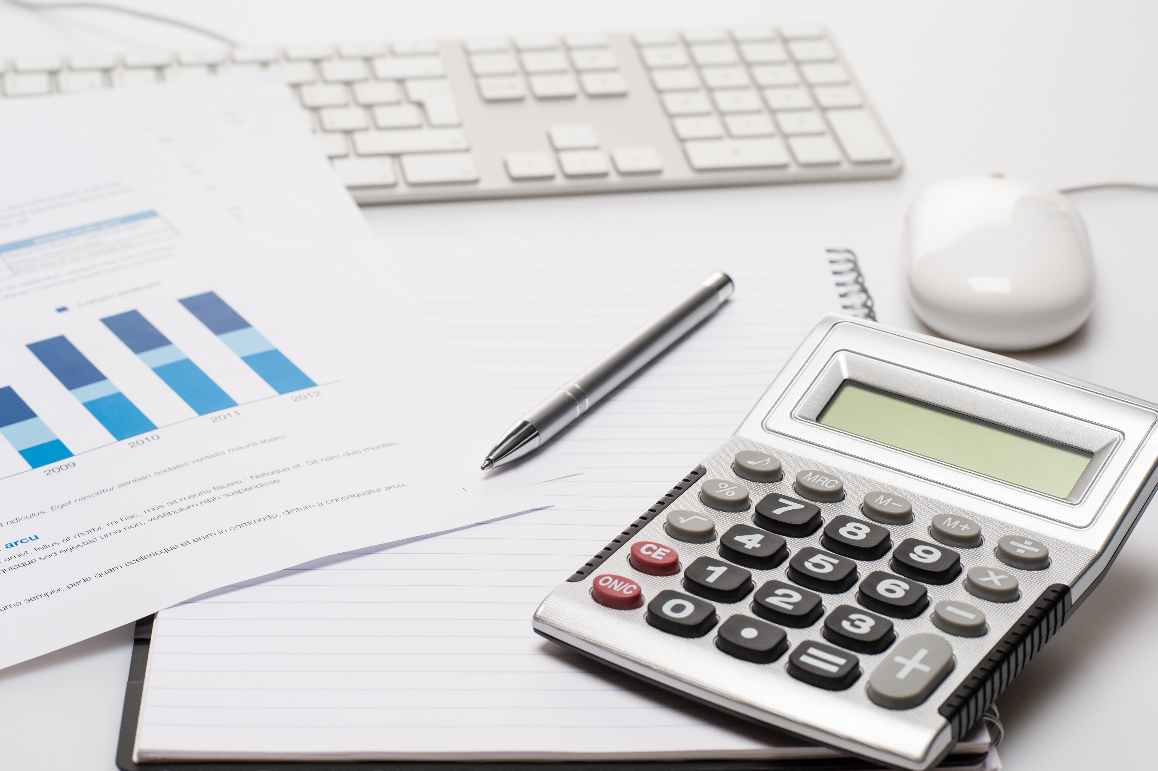 White office desk with supplies: calculator, pen, notepad, papers with graphs, keyboard, and mouse