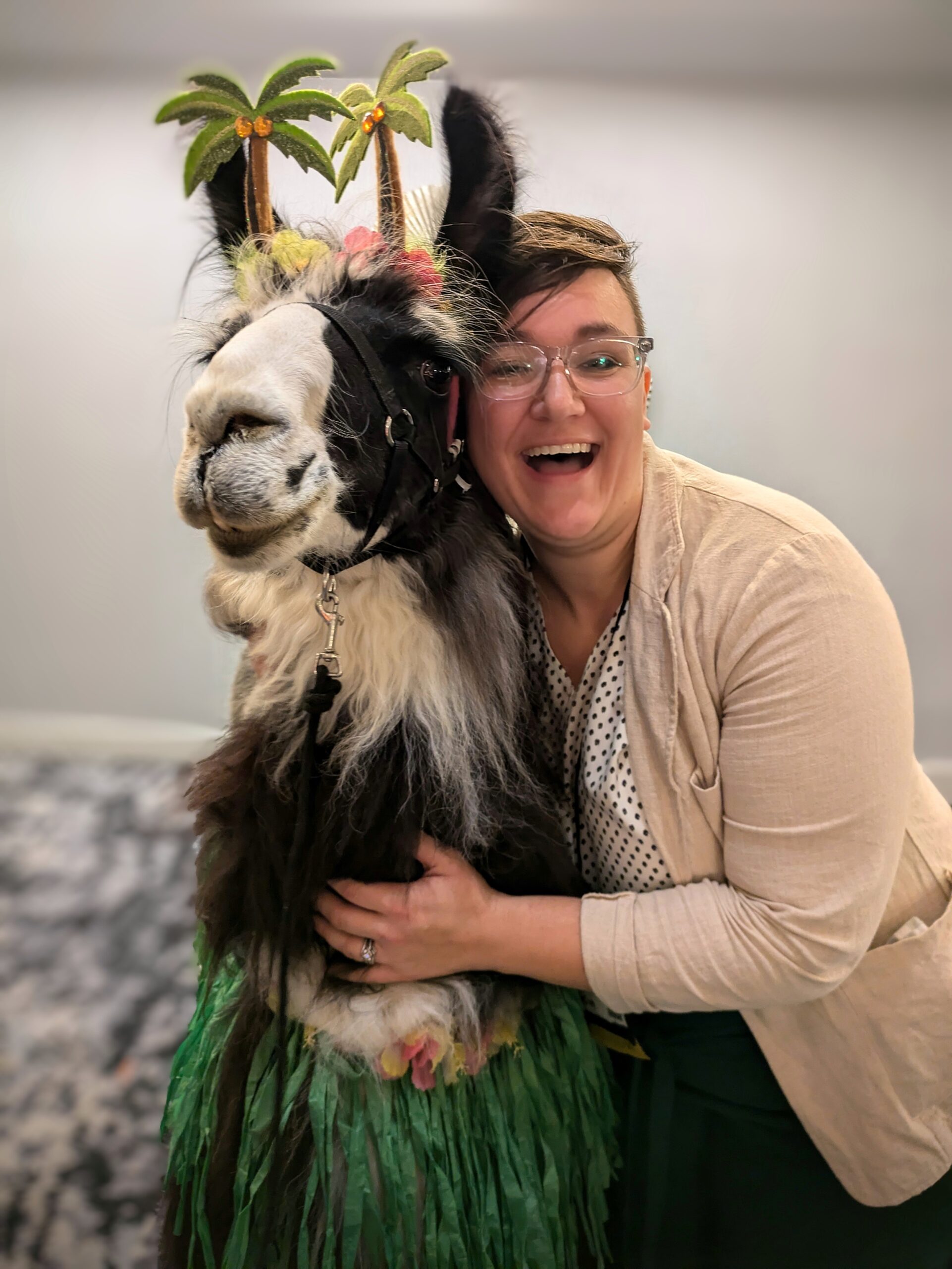 Woman with glasses and business attire smiling and hugging a llama. 