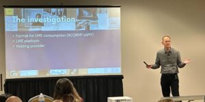 A presenter--Alan Pringle--stands in front of a projection screen that reads “The investigation” with bullet points below about LMS formats, platforms, and hosting providers. Several audience members sit in rows of chairs, listening attentively.