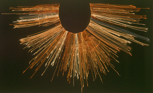 A quipu, an ancient Incan record-keeping device, photographed against a dark background. Dozens of knotted cords in shades of orange, tan, and cream fan outward from a central oval object, resembling a sunburst. The varying colors, lengths, and knotted patterns along each cord represent encoded numerical and categorical data — an early metadata system used by the Inca Empire to organize and store information centuries before digital cataloging existed."